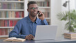 © stockbakers - Young African Man Talking on Phone while using Laptop in Office