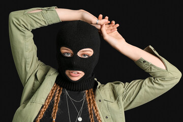 Wall Mural - Portrait of young woman in balaclava with hands above her head on black background