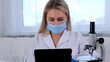 © Евгения Медведева - female research chemist scientist in a protective mask and gloves, dressed in a medical suit, with a tablet computer and a microscope, conducts research in the laboratory.