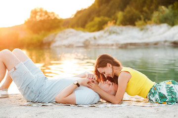 A young couple in love are resting on the shore of a lake, lying on the sand in summer sunny weather at sunset.