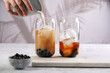 Female hand pouring milk into glass with black tea, ice cubes and cooked tapioca pearls for trendy bubble boba ice tea, two small grey ceramic bowls on marble board on grey concrete background