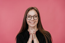 Close-up Portrait Of A Young Female Student With Glasses In A Black T-shirt, Who Puts Her Hands Together,  Looks At Camera With Gratitude, On A Pink Background. Italian Girl Happy With New Glasses.