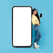 © Prostock-studio - Happy young woman standing near big cellphone with empty white screen, showing mobile device on blue background. Mockup