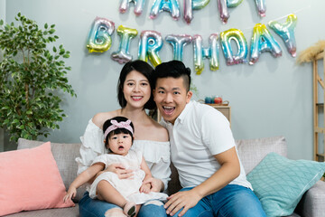 Wall Mural - sweet asian nuclear family with a lovely baby smiling at the camera while celebrating birthday in a modern bright living room with festive decoration at home