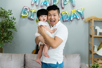 Wall Mural - front portrait of a cheerful asian father holding his innocent baby daughter and looking at the camera with smile on the occasion of birthday at home.