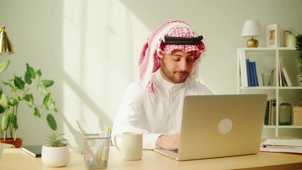Middle eastern man using laptop. Home office. Male person texting at computer keyboard in living room. Wearing traditional Islamic clothes. Communicating with family and friends online.
