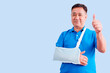 © krongthip - Old man with broken arm thumbs up on blue background.