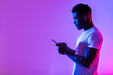 Side view of young black man in earphones listening to music, using new mobile app on smartphone in neon light, free space