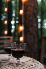 Two glasses of red wine standing on the wooden table with blurred trees and lights on the background.