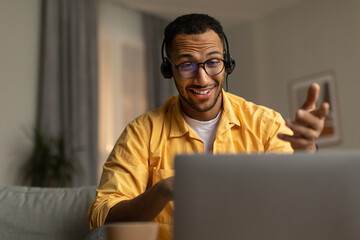 Millennial black man in headset having web call, using laptop computer, sitting on couch, talking at webcam at home