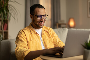 Wall Mural - Beautiful young black man working on laptop pc, enjoying remote job or education in living room