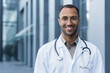 © Liubomir - Close-up photo portrait of young African American doctor outside hospital, man in medical coat smiling and looking at camera.