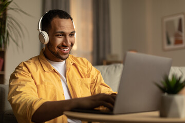 Wall Mural - Joyful young African American man using laptop, having remote job, wearing headphones at home