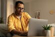 © Prostock-studio - Focused millennial black man in glasses using laptop, working online, having online business meeting at home office