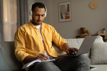 Wall Mural - Focused young black man in headphones having online business conference on laptop, taking notes at home