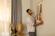 © Prostock-studio - Young black man putting picture frame, hanging painting on wall, empty space. Home interior and domestic decor