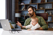 © YURII MASLAK - Father on maternity leave, holding an online conference, a meeting in the home office with a small child in his arms. Dad with a child. Freelancer combines child care and work. Paternity leave