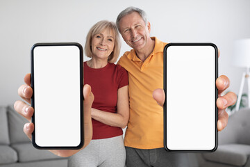Happy Senior Couple Standing At Home And Showing Blank Smartphones At Camera