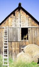 Old Wooden Barn With A Chair In An Upper Floor Window, Ladder Leaning Against The Building And Hay Bales In The Foreground