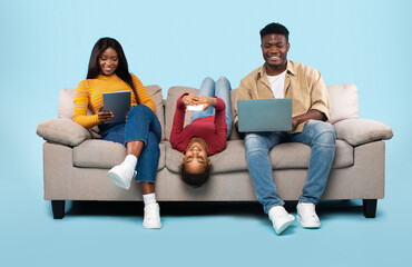 Gadgets addiction concept. African american family of three using electronic devices, sitting on sofa on blue background