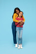 © Prostock-studio - Happy black mother hugging her lovely daughter standing in studio over blue background, full length shot