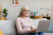 © Prostock-studio - Happy caucasian mature gray-haired female in glasses typing on laptop, watching video in living room interior