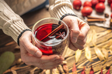 Hands of woman in a sweater are holding a glass cup with mulled wine on a wooden background with autumn leaves. Concept of a traditional warming beverage