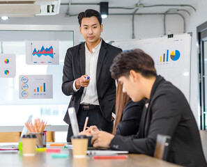 Asian handsome professional successful businessman presenter standing holding pen pointing presenting company graph chart paperwork document on glass board to male female colleagues in meeting room