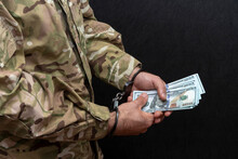A Soldier In A Military Camouflage Uniform Is Handcuffed, Holding A Bundle Of $100 Bills In His Hands On A Black Background. Concept: Bribery In The Army, Illegal Sale Of Weapons, Military Court.