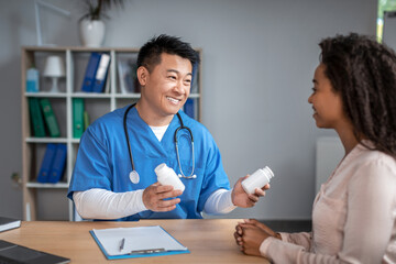 Happy young asian man doctor in uniform consults to black lady patient, offers jars with pills in office