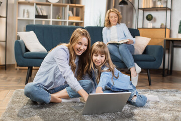 Canvas Print - Cute blond girls sisters sitting on warm floor play at home together using laptop, while happy mother relax on sofa on background.