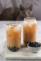 Two glasses with black tea, milk, ice cubes and cooked tapioca pearls for trendy bubble boba ice tea, two small grey ceramic bowls on marble board on pastel tropical background, cat in the back