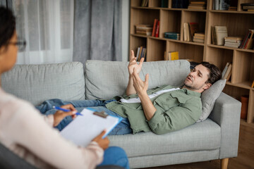 Wall Mural - Sad young european guy patient lies on sofa and talks with black woman doctor in office