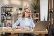 © sofiko14 - Portrait of beautiful confident with blonde hair and on headphones clever woman wearing casual clothes, sitting at table in front of computer and checking information in presentation on internet