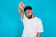 © Jihan - Discontent bearded caucasian man wearing white T-shirt over blue background shows disapproval sign, keeps thumb down, expresses dislike, frowns face in discontent. Negative feelings.