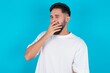 © Jihan - Sleepy bearded caucasian man wearing white T-shirt over blue background yawning with messy hair, feeling tired after sleepless night, yawning, covering mouth with palm.