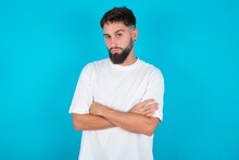 Bearded Caucasian Man Wearing White T-shirt Over Blue Background Frowning His Face In Displeasure, Keeping Arms Folded, Waiting For An Explanation.