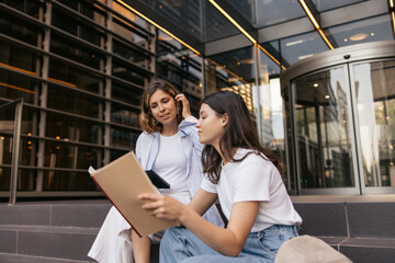 Successful young caucasian high school students check their homework before class sitting outside. Blonde with brunette wear light spring clothes. Study concept