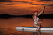 Junge Frau im Badeanzug sitzt auf einem SUP und paddelt beim Sonnenuntergang am See