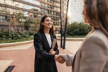 Wall Mural - Cheerful young caucasian female director shaking hands with job seeker spending time outdoors. Brunette wears black jacket to interview. Management concept 