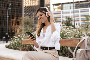 Wall Mural - Cheerful young caucasian girl listens to music using headphones and smartphone sitting on bench. Blonde wears classic style of clothing. Audio technology concept