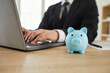 © Studio Romantic - Close up of a blue piggy bank on the office desk of a businessman working on a modern laptop computer in the background. Business, finance, investment, profit concepts