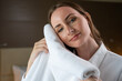 Happy woman in white shirt smells soft terry towel on blurred background. Female guest enjoys facilities of hotel standing in room against lamp closeup