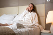 © Grustock - Woman rests on king-size bed calling receptionist to order dinner to hotel room. Brown-haired tourist wearing terry bathrobe enjoys vacation at resort