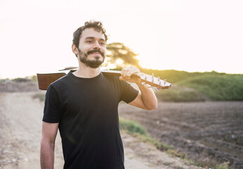 Portrait of young musician with his guitar on his shoulder on the road at sunset