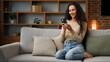Smiling Hispanic Caucasian 30s woman housewife girl sitting on couch cozy sofa at home using smartphone for ordering food delivery chatting with mobile app looking at phone screen with smile browsing