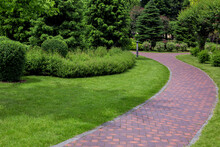 Curved Pathway Paved Brick Stone Tiles In Park Among Plants, Foliage Bushes And Pine Trees Surrounded By Eco Friendly Green Lawn On Sunny Day And Iron Ground Lantern Garden Lighting, Nobody.