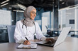 © Liubomir - Arab muslim woman in hijab doctor working in modern clinic office with laptop, doctor on paper work in white medical coat with stethoscope and glasses.