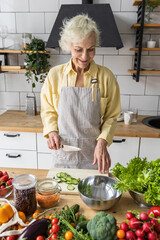 Wall Mural - Attractive senior woman with grey hair cooking healthy food on her kitchen at home. Mature female preparing fresh vegetarian salad with organic ingredients from the market: tomato, cucumber, pepper