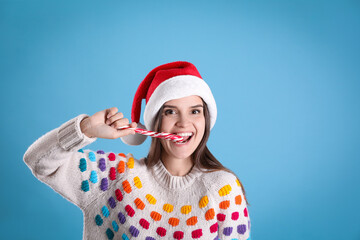 Poster - Pretty woman in Santa hat and festive sweater eating candy cane on light blue background